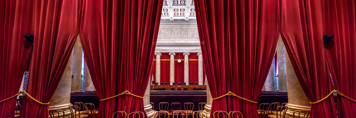 Photograph of an ornate courtroom with heavy red curtains, marble columns and chairs.