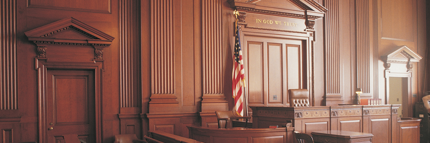 Photograph of a dark wood-panelled courtroom with an American flag and a judge's bench above which the words "In God We Trust" are written in gold letters.