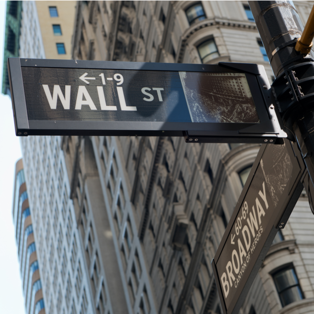 A photograph of a street sign in New York City with intersection signs for "Wall St." and "Broadway."