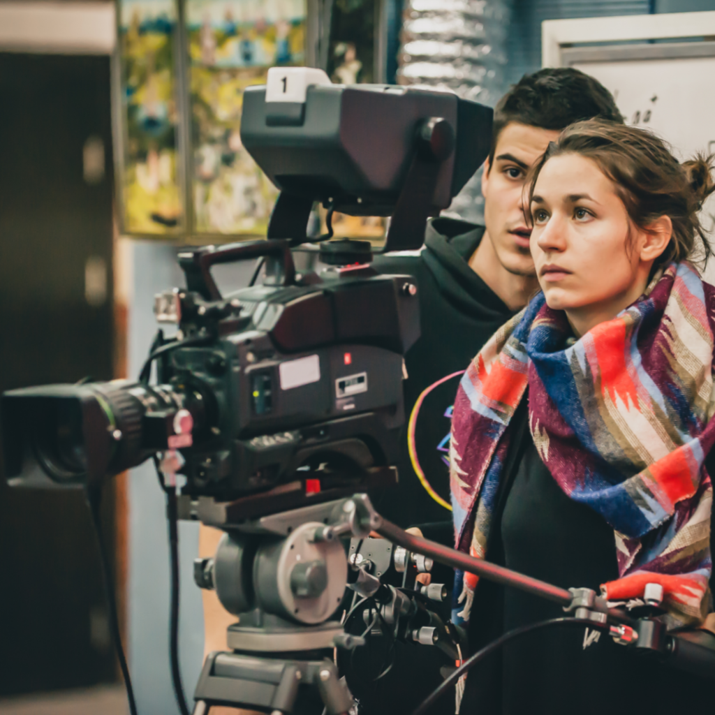 Photograph of a man wearing a black hoodie and a woman wearing a colorful scarf, looking through a large movie camera.