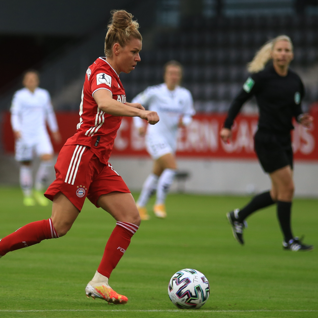 Female soccer player dressed in red, about to kick a multicolored soccer ball, watched by referee and other players.