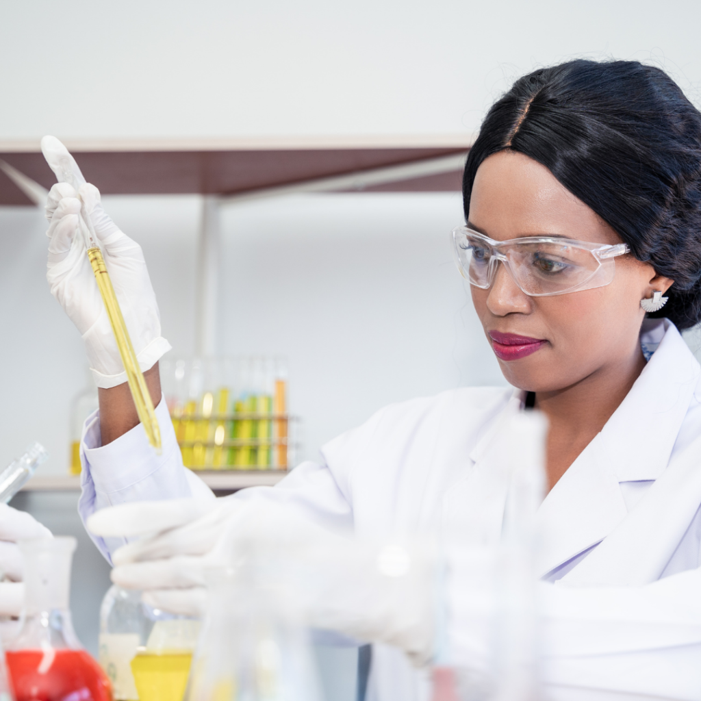 Photograph of a female chemist wearing safety glasses and a white lab coat, using a pipette to put yellow liquid into a glass vial.