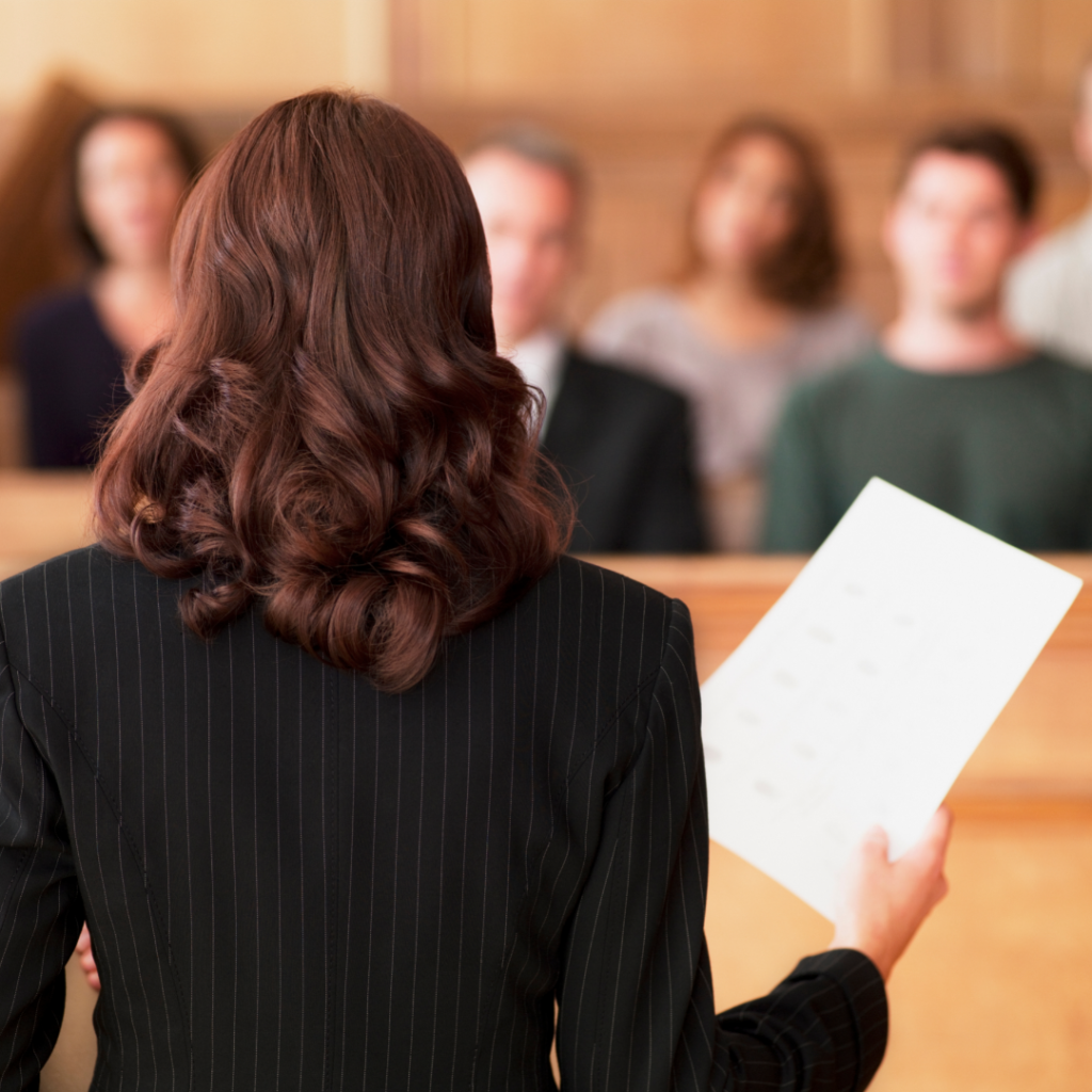 Photograph showing the back of a female lawyer holding a document and speaking to a jury.