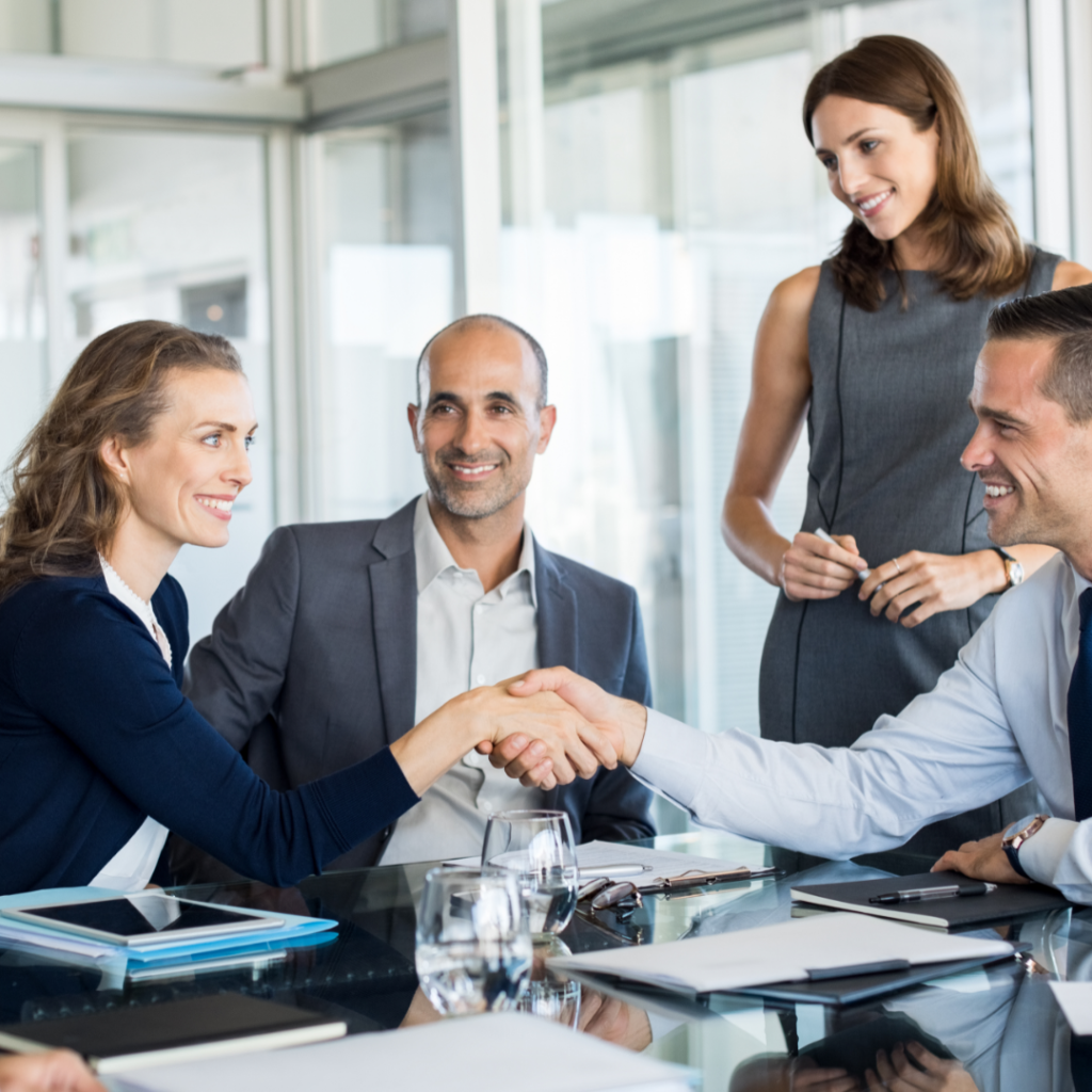 Photograph of male and female business people shaking hands with other business people looking on.