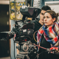 Media, Entertainment & Fine Arts Photograph of a man wearing a black hoodie and a woman wearing a colorful scarf, looking through a large movie camera.