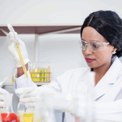 Pharma, Healthcare & Life Sciences Photograph of a female chemist wearing safety glasses and a white lab coat, using a pipette to put yellow liquid into a glass vial.
