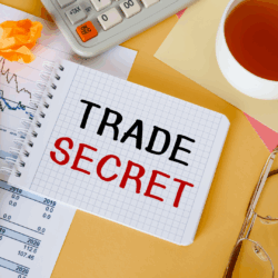 Trade Secret Litigation Photograph of a desktop with a calculator, a cup of tea and a small spiral notebook with the words "Trade Secret" written in capital letters.